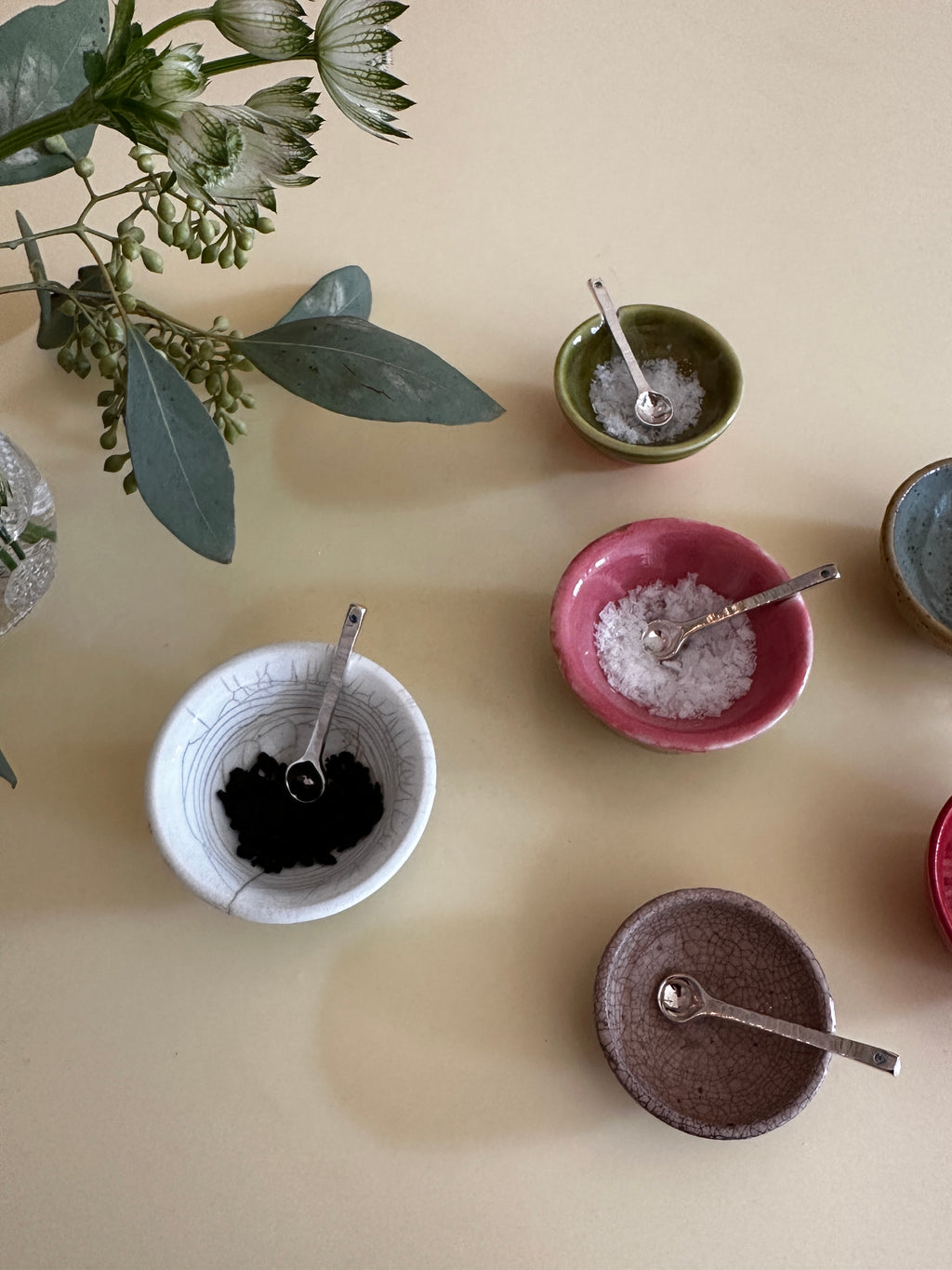 Collection of small ceramic bowls with spoons on a beige surface, accompanied by green leaves.